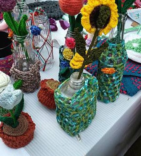 Crochet flowers and accessories displayed on a white tablecloth at a creative market stall. Colorful, handcrafted flowers in glass bottles feature various designs, including sunflowers and roses. Beside them are crocheted pouches, coasters, and bags in vibrant hues. The setup includes items with intricate patterns, showcasing craftsmanship. The background displays a mix of textures and colors, highlighting the diversity of crocheted items available.